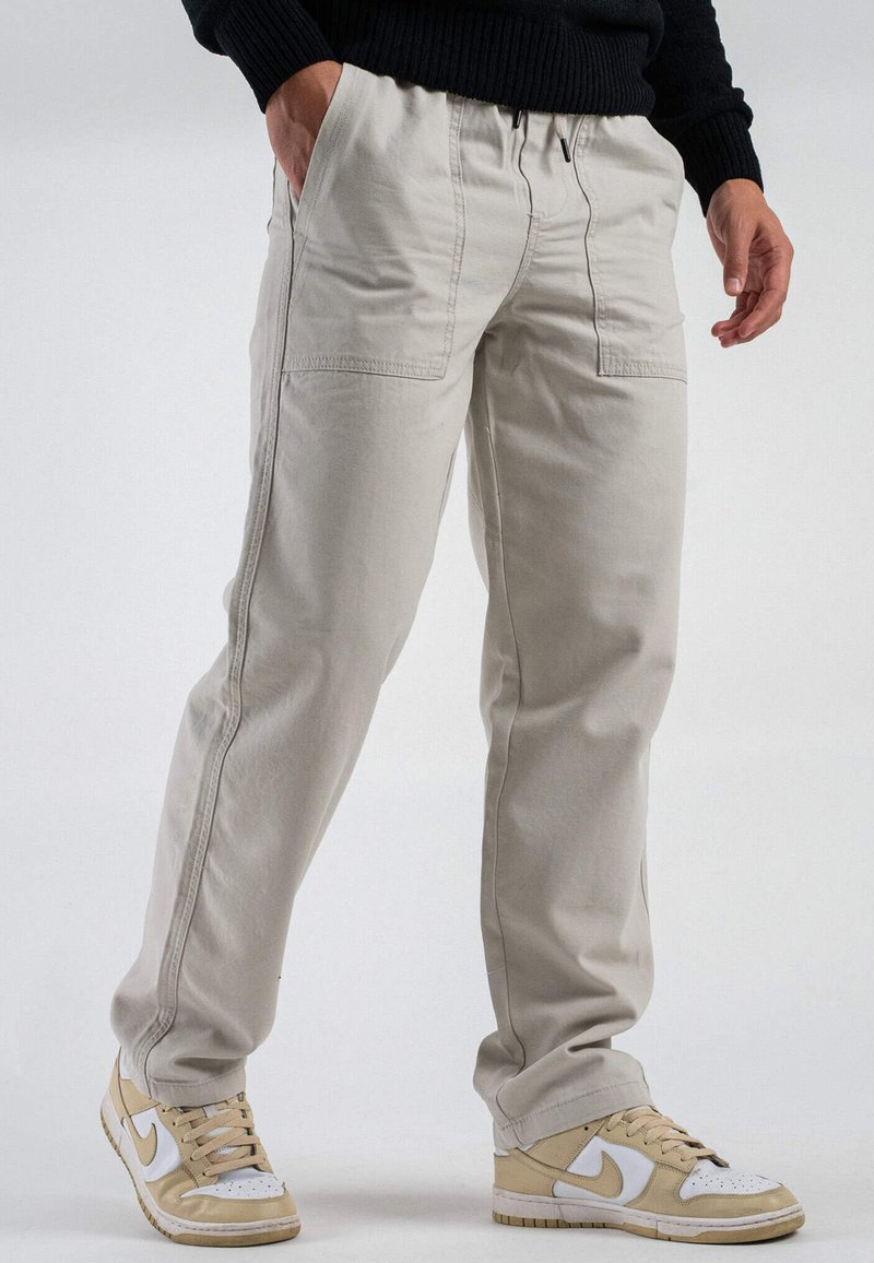 Pantaloni cargo beige chiaro con vita elastica, multiple tasche e una vestibilità rilassata, abbinati a sneakers beige e bianche.