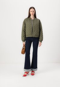 Veste bomber matelassée vert olive avec une taille en bord-côte ajustée, associée à un jean foncé évasé et des chaussures à talons rouges, portant un sac à main marron.