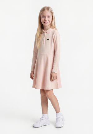 Niña joven con cabello largo rubio, que lleva un vestido rosa claro de manga larga y zapatillas blancas, sonriendo y de pie frente a un fondo blanco.