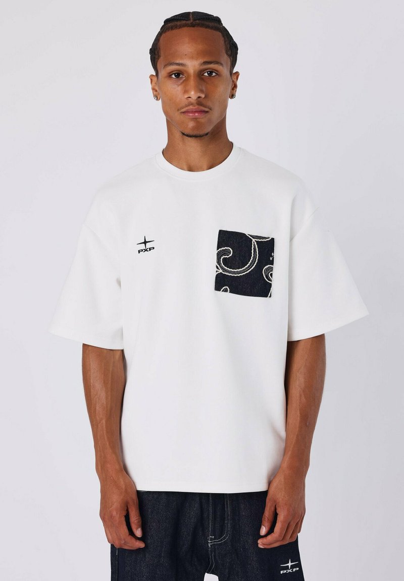 Camiseta blanca de manga corta con un bolsillo negro decorado, con un logo en el lado izquierdo. La tela parece suave y ligera.