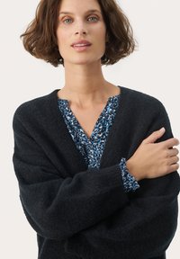Maglione nero strutturato sovrapposto a una blusa blu con motivo floreale. La blusa presenta polsini a balza e un collo con apertura parziale.