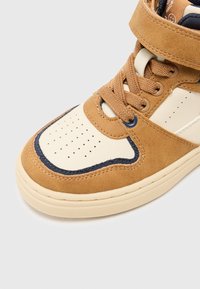 Brązowe i beżowe zamszowe sneakersy z kremowym noskiem, granatowymi akcentami, perforowanymi wzorami oraz wyściełanym paskiem wokół kostki dla wsparcia.