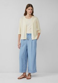 Cardigan leggero giallo chiaro sopra una maglietta aderente color crema, abbinato a pantaloni larghi azzurri chiari con vita elastica e dettagli a nappina.