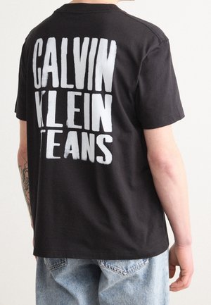 Osoba w czarnej koszulce z dużym białym napisem "CALVIN KLEIN JEANS" na plecach, w połączeniu z jasnoniebieskimi dżinsami.