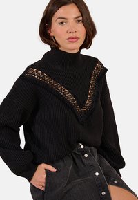 Maglione nero lavorato a maglia con colletto alto, caratterizzato da un design a scollo a V con motivo in pizzo. Texture morbida, abbinato a una gonna nera con bottoni.