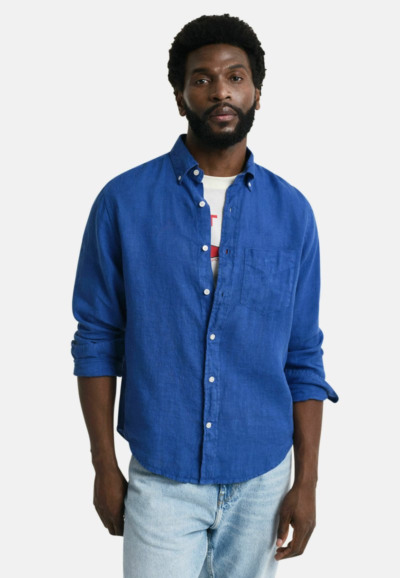Camisa de lino azul con botones, con un bolsillo en el pecho, mangas remangadas y un corte relajado. Llevada sobre una camiseta gráfica blanca y unos jeans claros.