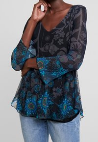 Mujer con una blusa negra transparente con patrones florales azules y verdes y mangas acampanadas, combinada con jeans de un azul claro.