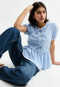 Ljusblå och vit rutmönstrad blus med puffärmar, samlad midja och rosettaccent. Bärs med blå denimjeans.