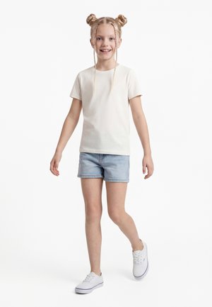 Usměvavá dívka s blond vlasy učesanými do dvou drdolů a copů, oblečená v bílém tričku, džínových šortkách a bílých teniskách, kráčející vpřed.