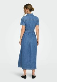 Femme debout portant une robe midi en jean bleu à manches courtes avec une ceinture et des chaussures plates noires, vue de dos.