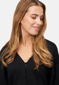 Czarna bluzka z dekoltem w serek, zapinana na okrągłe guziki z przodu. Miękki materiał o gładkiej fakturze i luźnym kroju. Długie włosy uczesane rozpuszczone.