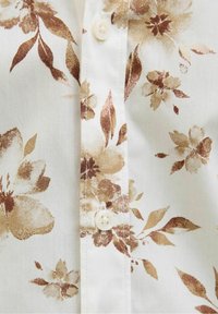 Bílá látka s hnědým květinovým vzorem, obsahující okvětní lístky a listy; součástí je zapínání na knoflík a viditelné šicí detaily.