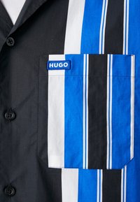 Close-up van een zwart shirt met een verticale gestreepte zak in blauw, wit en zwart, met een blauwe "HUGO" label op de borst.