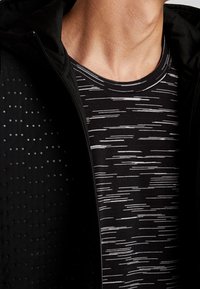 Chaqueta atlética negra con textura de puntos, desabrochada sobre una camiseta a rayas blanca y negra. Primer plano del escote y la parte superior del pecho.
