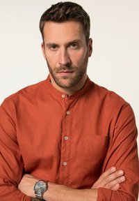Rostfarbendes Hemd mit Mandarin-Kragen, Brusttasche und glatter Textur. Das Modell trägt eine silberne Uhr am Handgelenk.