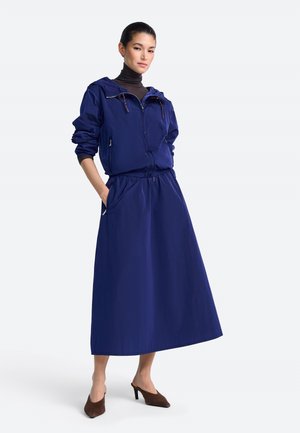 Mujer con chaqueta con capucha azul marino y falda larga a juego, con zapatos marrones de tacón alto, de pie con las manos en los bolsillos.