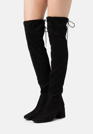 Botas de ante negras por encima de la rodilla con tacón cuadrado, que presentan detalles de lazo en la parte superior y una textura suave. Diseño de punta afilada.