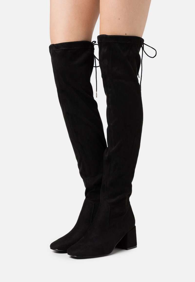 Botas de ante negras por encima de la rodilla con tacón cuadrado, que presentan detalles de lazo en la parte superior y una textura suave. Diseño de punta afilada.