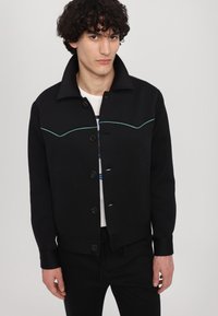 Černá bunda se strukturovaným límcem, zdobená tyrkysovým prošíváním na hrudi, zapínáním na knoflíky a bočními kapsami. Hladká tkanina.