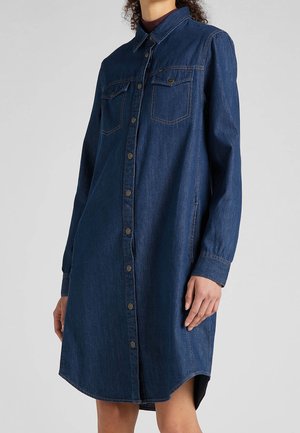 Vrouw draagt een donkerblauwe denim blousejurk met lange mouwen, dichtgeknoopt aan de voorkant met twee borstzakken en zijnaadzakken.