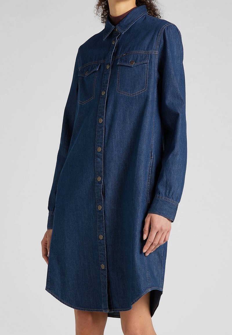 Femme portant une robe-chemise en denim bleu foncé à manches longues, boutonnée sur le devant, avec deux poches poitrine et des poches latérales.