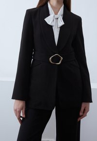 Zwarte getailleerde blazer met een smalle riem met een gouden ovale gesp, gecombineerd met een witte blouse met strik. Gladde stof en gestructureerde vorm.