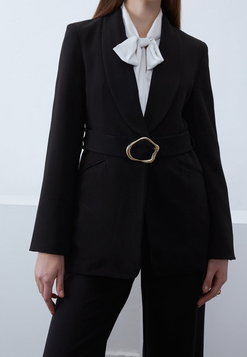 Zwarte getailleerde blazer met een smalle riem met een gouden ovale gesp, gecombineerd met een witte blouse met strik. Gladde stof en gestructureerde vorm.