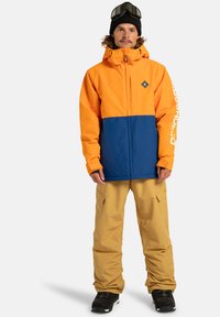 Mężczyzna ubrany w pomarańczową i niebieską zimową kurtkę, beżowe spodnie narciarskie, czapkę beanie w kolorze czarnym i czarne buty, stojący na białym tle.