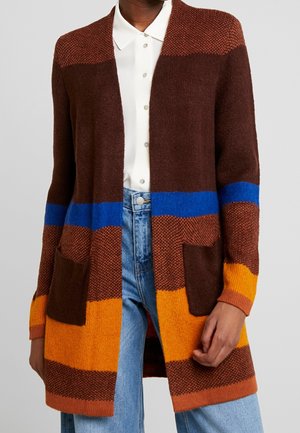 Personne portant un jean bleu, une chemise blanche boutonnée et un long cardigan tricoté multicolore avec des rayures marron, orange et bleues et des poches avant.