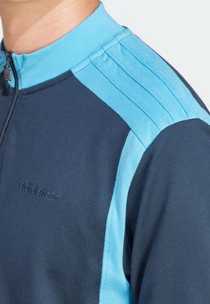 Navy blue athletic jacket with a high collar, featuring light blue shoulder stripes and a small embossed adidas logo on the chest.