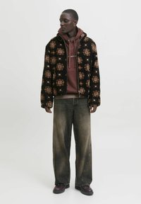 Giacca in pile nera con dettagli a fantasia, sovrapposta a una felpa bordeaux, abbinata a jeans larghi e sbiaditi grigi e scarpe marroni.