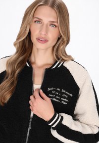 Sort og creme bomberjakke lavet af tekstureret fleece, med ribbede manschetter med sorte og hvide striber, samt broderet tekst foran.