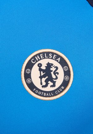 Un tissu bleu à texture lisse présente un logo circulaire du Chelsea Football Club en blanc et noir, avec un design de lion et des éléments décoratifs.