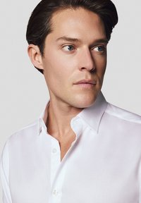 Biała koszula z odkrytym kołnierzykiem, charakteryzująca się gładką teksturą i delikatnym połyskiem, noszona przez osobę na neutralnym tle.