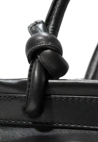 Detail einer schwarzen Lederhandtasche, die einen knotenförmigen Griff, eine glatte Textur und präzise Nähte entlang der Kante zeigt.