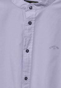 Jasnofioletowa koszula z długim rękawem i stójką, z ciemnymi guzikami i małym nadrukowanym logo po lewej stronie. Gładka faktura materiału.