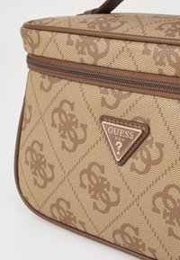 Borsa a tracolla marrone con motivo, realizzata in tessuto canvas texturizzato, con chiusura a zip e un prominente logo triangolare Guess sulla parte anteriore.