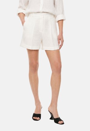 Persona che indossa pantaloncini di lino bianchi, camicia bianca a maniche lunghe con polsini arrotolati e sandali neri con tacco aperti in punta, le mani nelle tasche.