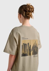 Beige bomulls-T-shirt med grafiskt tryck av berg och stadssilhuett på baksidan, med svart textdetaljer. Avslappnad, oversize passform.