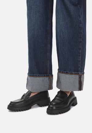 Czarne skórzane mokasyny z masywną podeszwą, z przeszywanym paskiem na górze. Noszone z podwiniętymi, ciemnymi jeansami.