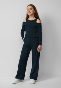 Navyblaues Langarm-Oberteil mit kalten Schultern, kombiniert mit lockeren navyblauen Hosen. Das Model trägt weiße Sneakers und steht vor einem neutralen Hintergrund.