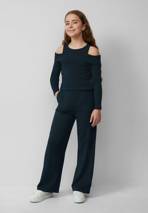 Marineblauw langemouwen top met koude schouders, gecombineerd met losse marineblauwe broeken. Model draagt witte sneakers en staat voor een neutrale achtergrond.