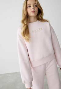 Rosa sweatshirt med ett guldtryckt "BAKER"-logotyp över bröstet. Har ribbade ärmslut och nederkant, tillverkad av mjukt, strukturerat material.