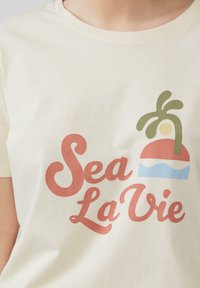 Hellbeige Baumwoll-T-Shirt mit stilisiertem "Sea La Vie"-Text in Koralle und Grün, sowie einer Strand-Szene-Illustration in Rot, Gelb und Blau.