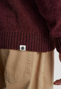 Burgunderfarbener Pullover mit einer strukturierten Strickmuster und geripptem Saum, mit einem kleinen Markenschild. Kombiniert mit hellbeigen Hosen.