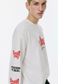 Junger Mann, der ein weißes langärmliges Hemd mit rotem "Stranger Things" und schwarzem "Monsters Are Real"-Text auf dem Ärmel trägt, vor einem schlichten Hintergrund.