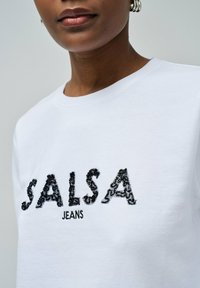 Sudadera blanca con un logotipo bordado texturizado en negro que dice "SALSA JEANS". La tela parece suave con un diseño clásico de cuello redondo.
