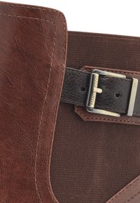 Botte de cheville en cuir marron avec un panneau en tissu texturé, une sangle noire ajustable avec une boucle en argent et des détails de couture visibles.