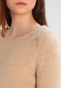 Beżowy sweter z dzianiny o szerokim dekolcie i miękkiej, puszystej teksturze. Posiada ściągacz na dekolcie i lekko przezroczystą tkaninę.