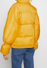 Giacca a vento gialla con una texture lucida, forma oversize e polsini elasticizzati. Presenta sezioni trapuntate e un design con colletto alto.